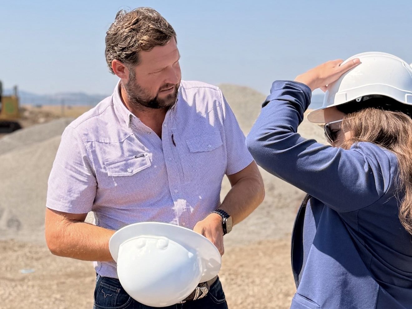 Two people in hard hats on a graded construction site, one holding a hard hat